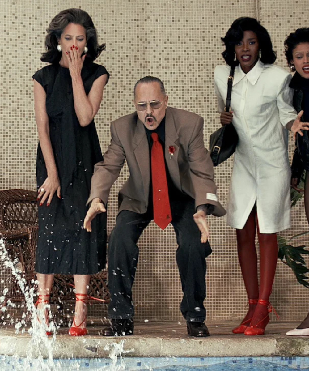 Willy à beira da piscina, com terno bege e gravata vermelha entre vestidos preto e branco elegantes