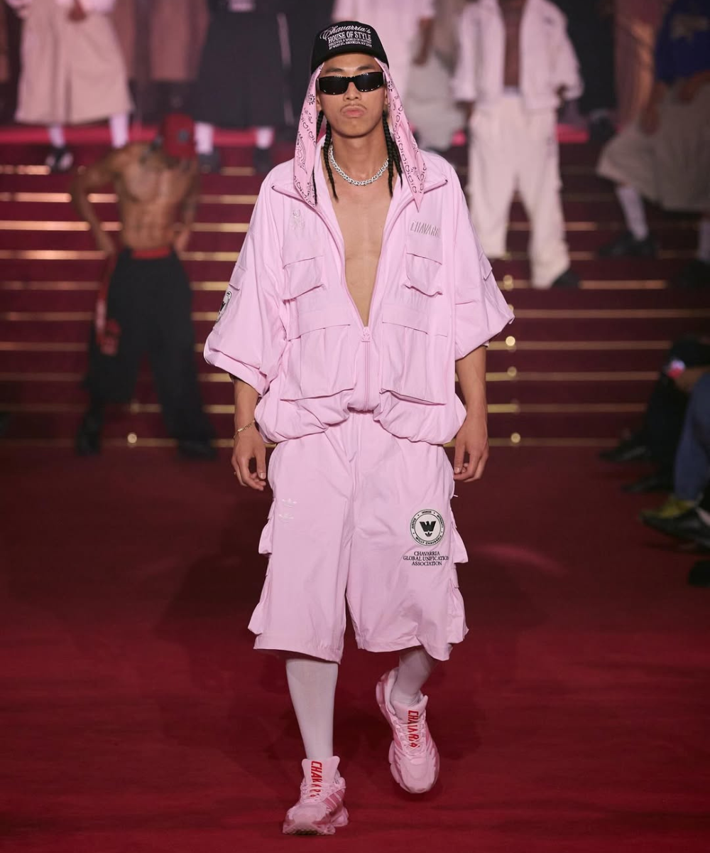 Willy na passarela: modelo desfila com conjunto rosa amplo, boné e óculos escuros, em clima urbano confiante.
