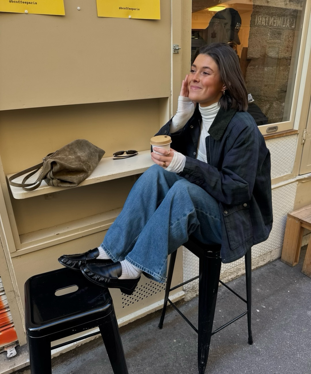 Looks de outono com jaqueta escura, gola alta e jeans amplos; modelo posa com café em clima acolhedor.