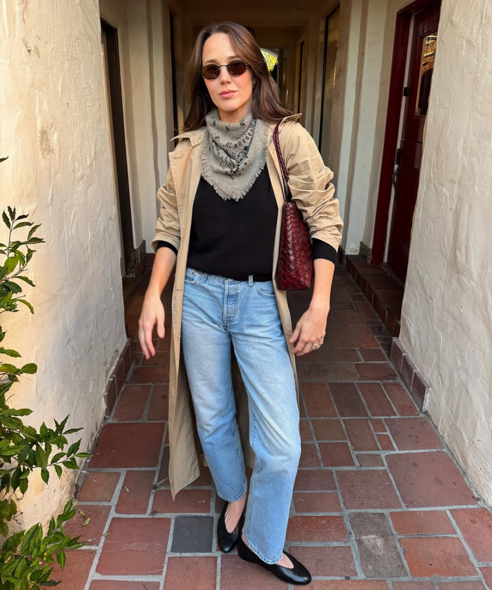 Looks de outono com modelo posa de trench bege, jeans reto, lenço cinza e bolsa vinho em corredor acolhedor.