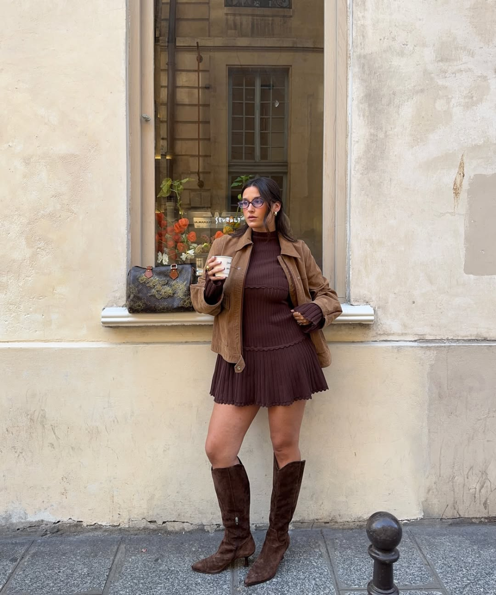 Looks de outono com modelo posa em vestido canelado marrom, jaqueta camel e bota de camurça diante de fachada clássica.