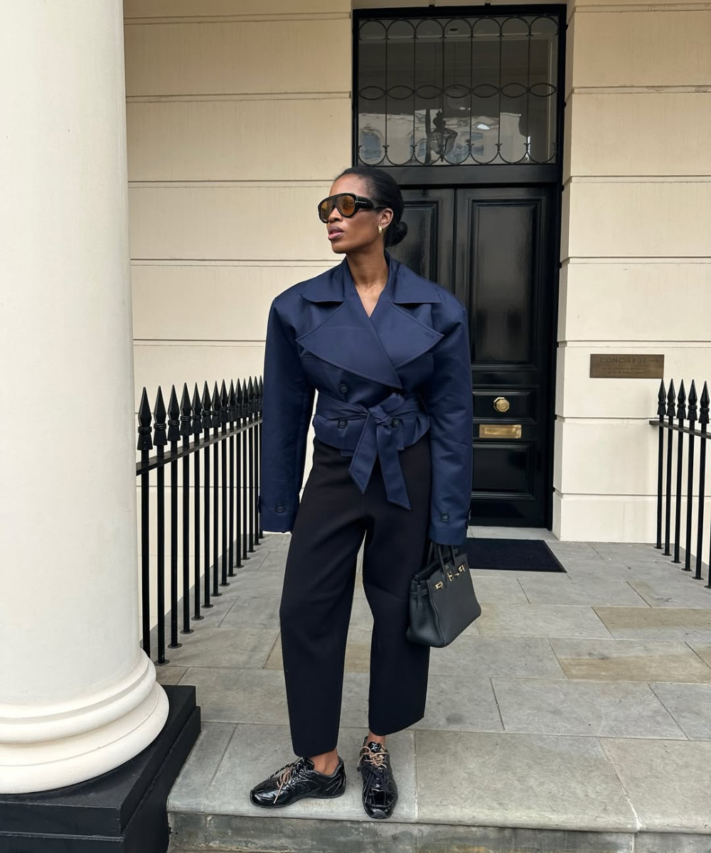 Looks de outono com modelo posa em trench curto azul-marinho, calça reta preta e óculos escuros em cenário elegante.