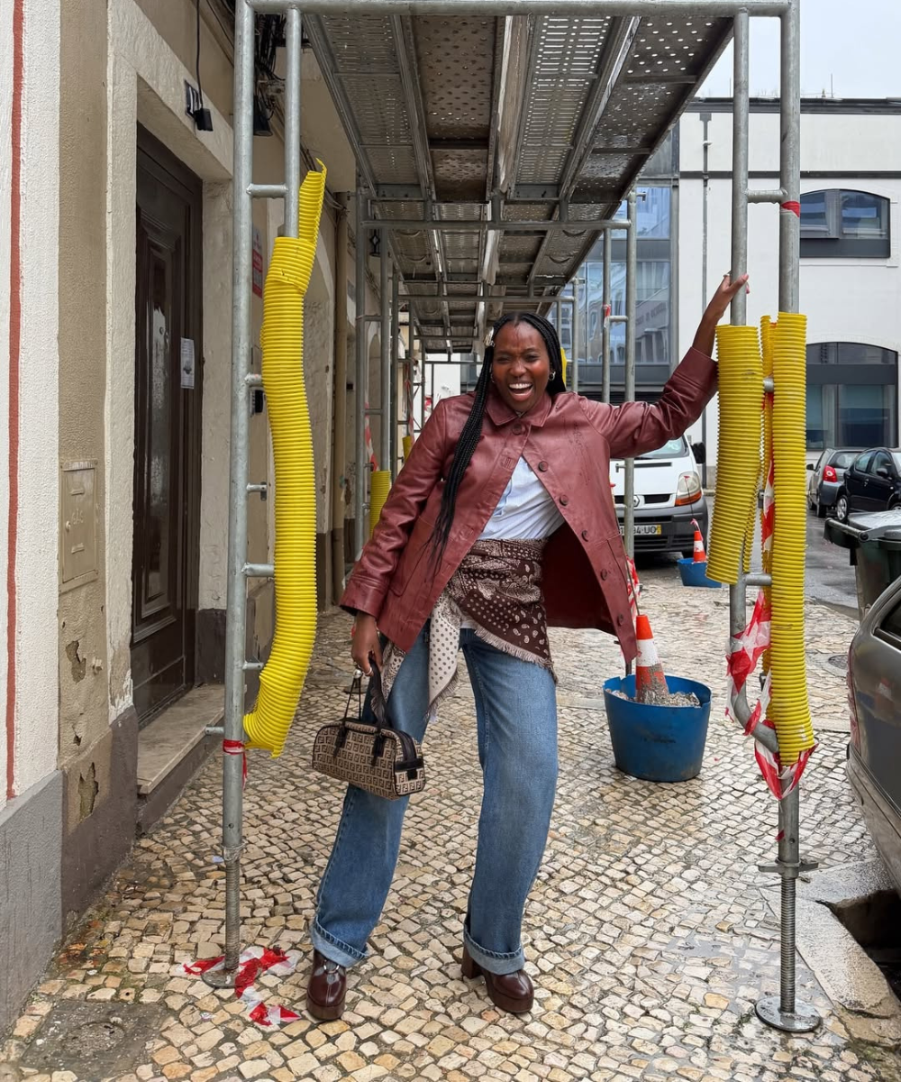 Looks de outono com jaqueta marrom, jeans amplo e lenço; modelo posa sorrindo na calçada, com energia urbana.