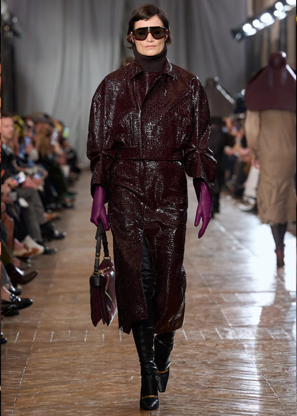 Outono/Inverno 2026: modelo desfila com casaco vinho texturizado, luvas roxas e botas pretas em clima sofisticado.