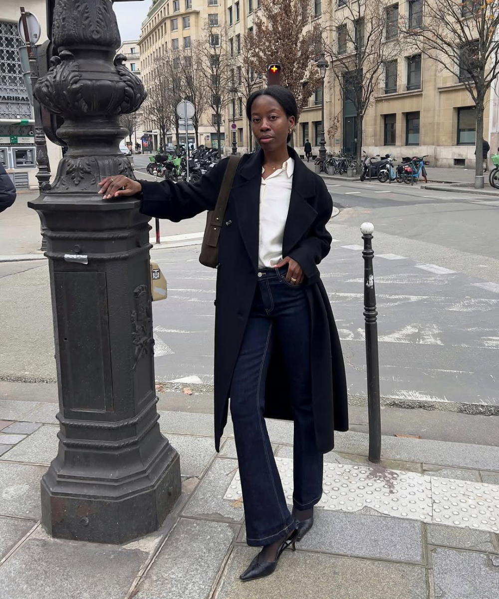 Look para trabalhar com calça jeans: modelo posa com calça jeans escura, camisa branca e sobretudo preto em rua urbana