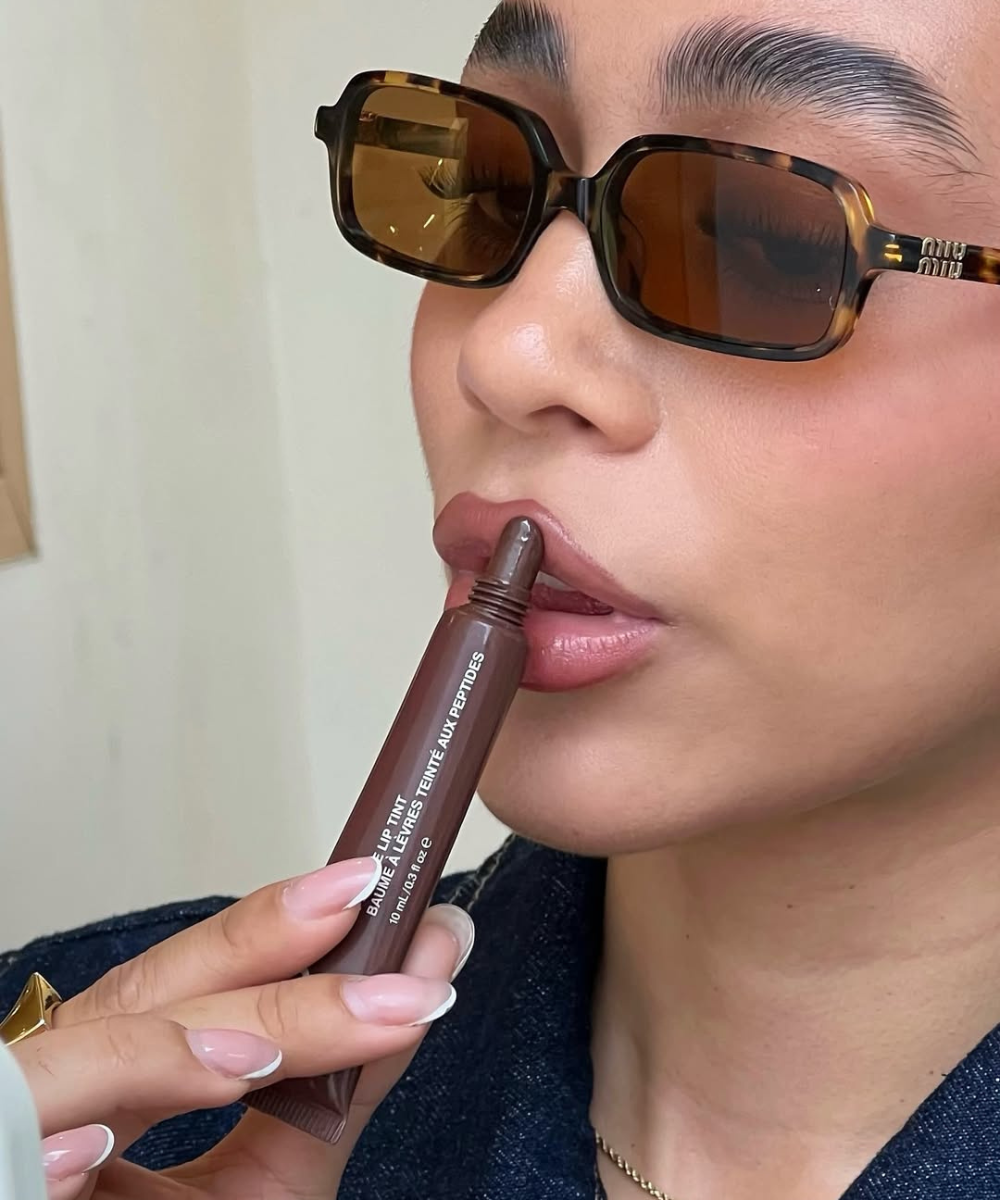 Modelo posa com gloss de chocolate nos lábios, óculos tartaruga e blazer escuro.