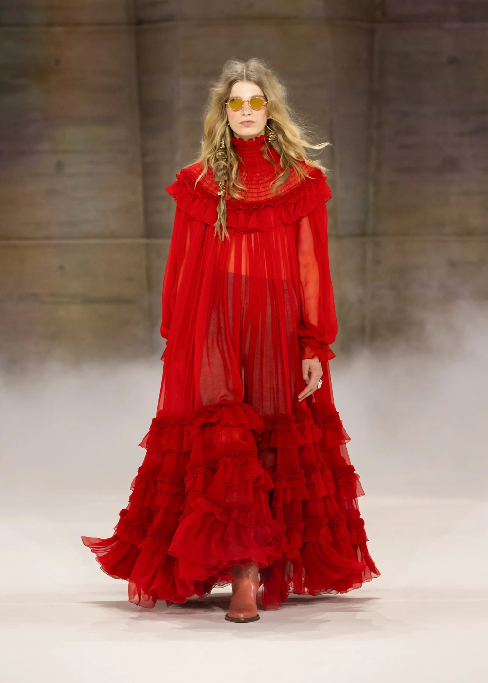 Outono/Inverno 2026: modelo desfila com vestido vermelho fluido de babados e transparência, em clima dramático.