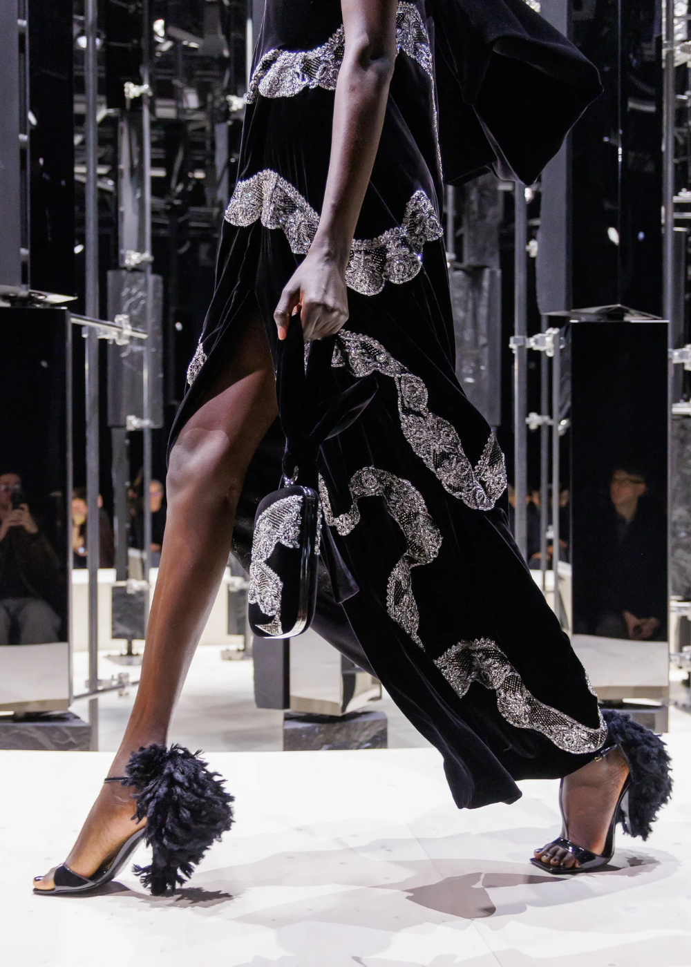 Outono/Inverno 2026: modelo desfila vestido preto de veludo bordado em prata, fenda alta e sandálias com plumas dramáticas