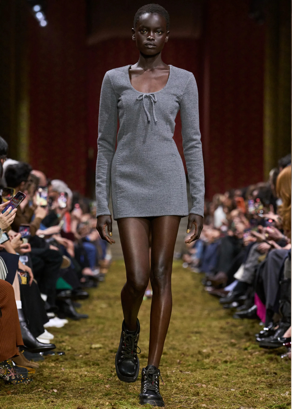 Outono/Inverno 2026: modelo desfila com vestido curto cinza canelado e coturnos pretos em passarela de clima sóbrio