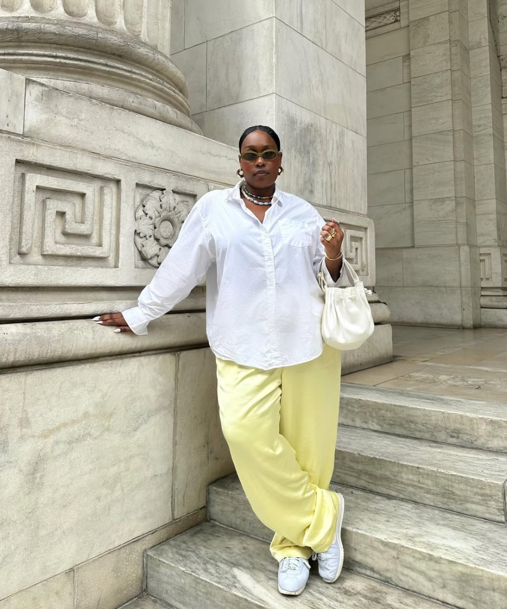 Modelo posa com calça pantalona amarela, tênis brancos e camisa ampla branca.