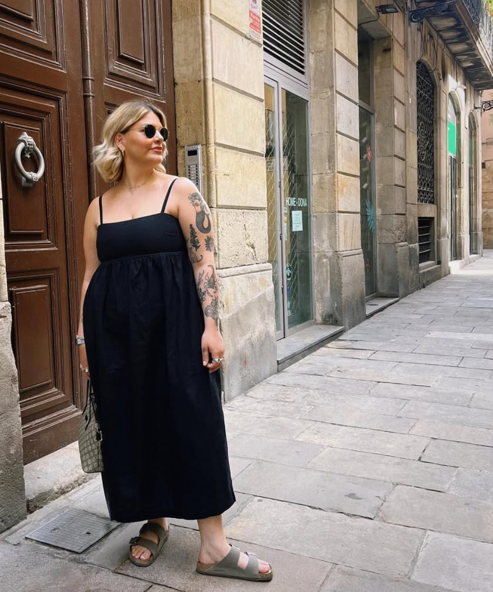 Que tipo de sapato usar com vestido soltinho: modelo posa com vestido preto amplo e rasteiras verdes em rua de pedra.