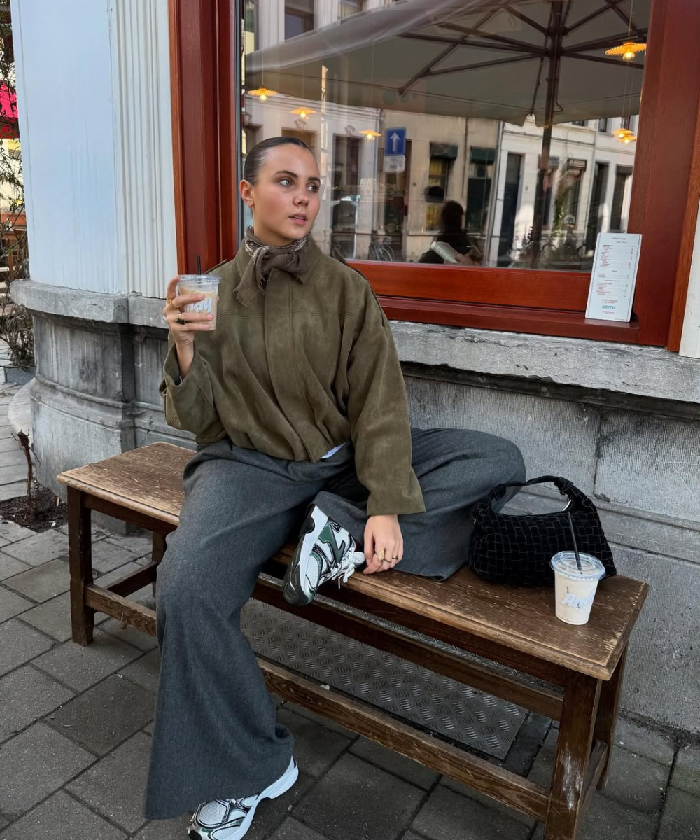 Modelo posa em banco com calça pantalona cinza e tênis esportivo, blusa verde de camurça; clima urbano sereno.