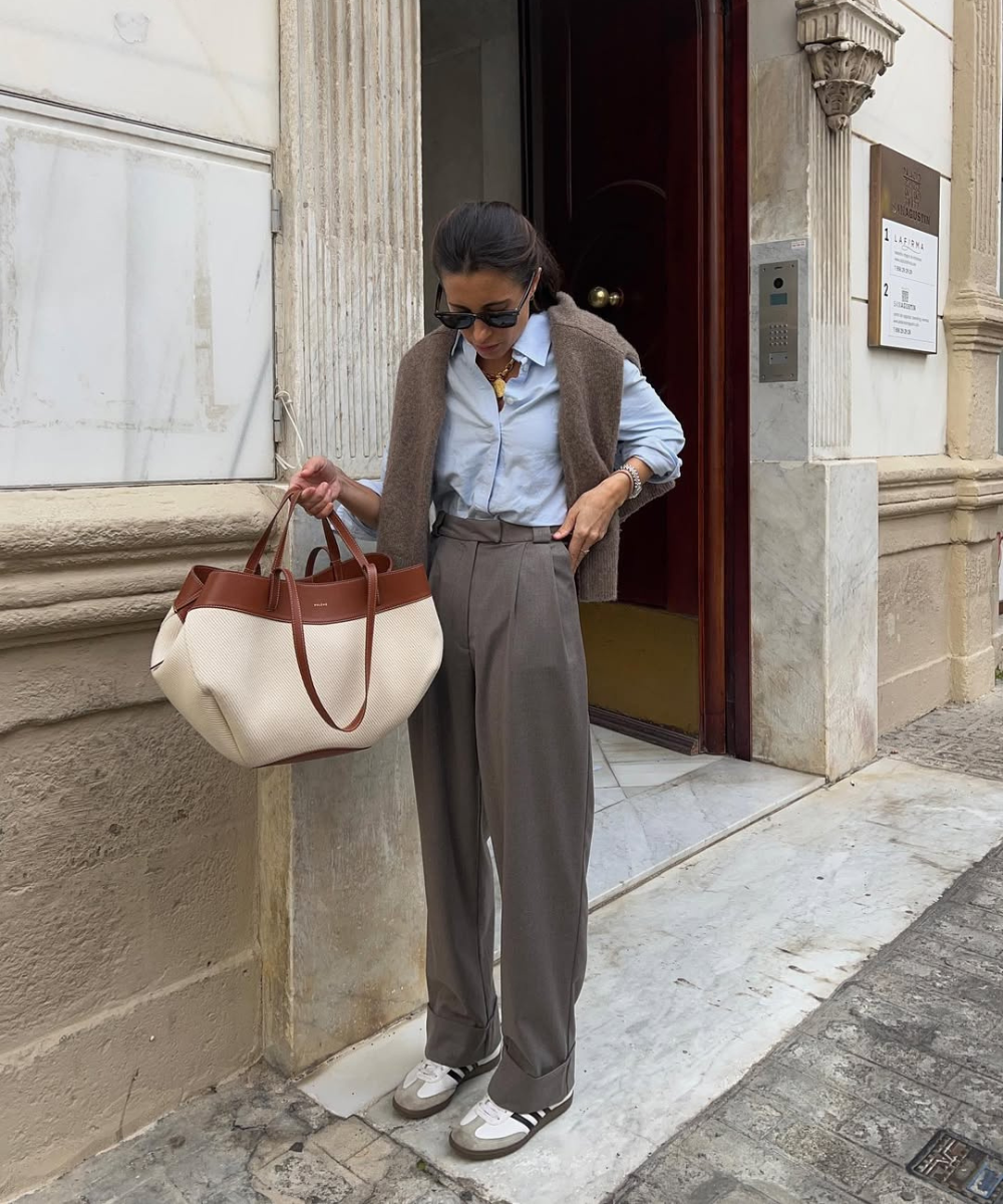 Modelo posa com calça pantalona cinza e tênis branco, camisa azul e tricô marrom.