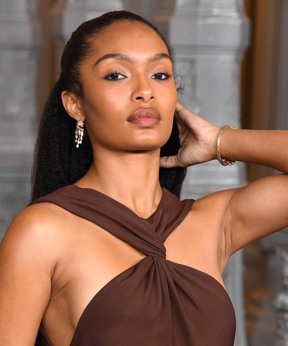 Penteados para casamento com cabelo polido e solto; modelo posa com vestido marrom drapeado e joias douradas elegantes