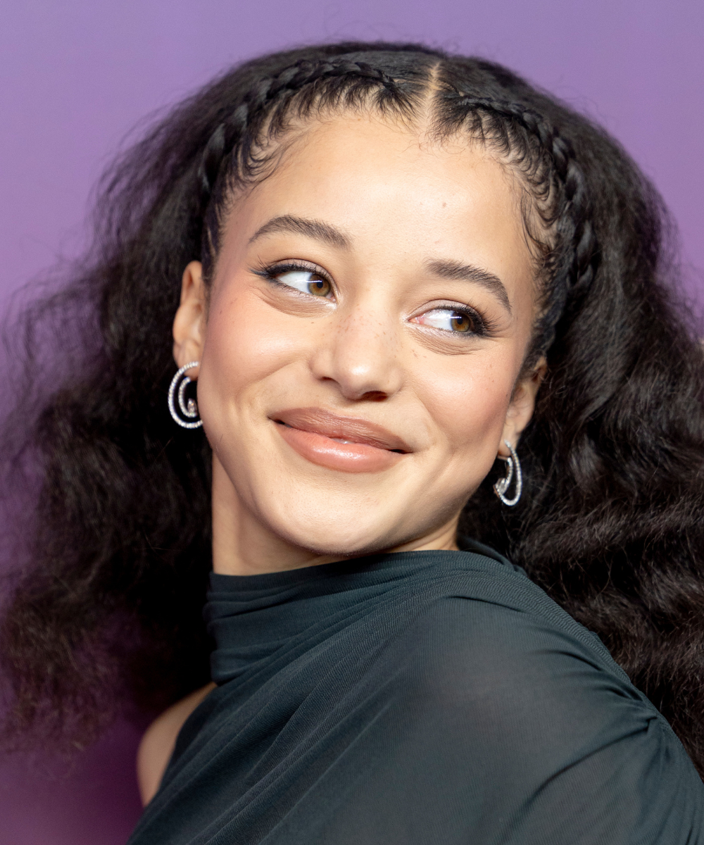 Penteados para casamento: modelo posa com tranças frontais e cabelo volumoso, vestido drapeado escuro e fundo lilás suave.