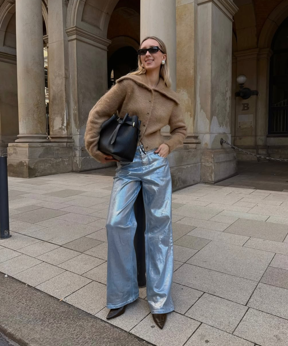 Modelo posa com calça resinada prata ampla, tricô marrom e bolsa preta diante de colunas, em clima elegante.