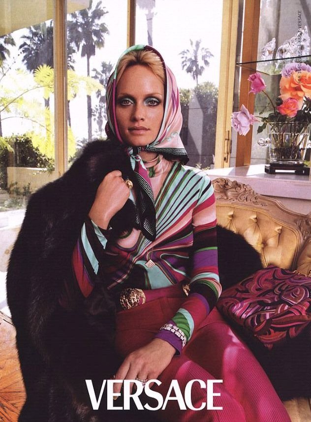 Modelo posa sentada com blusa listrada colorida, lenço na cabeça e casaco preto de pele, em ambiente sofisticado, estilo