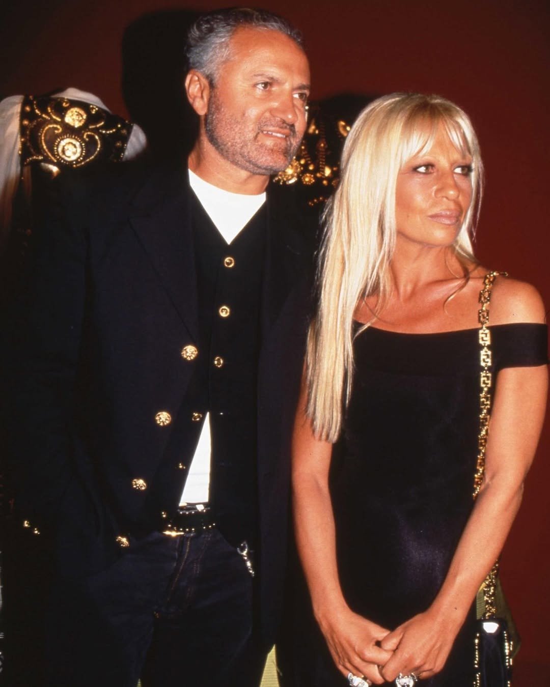 Casal posa elegante com peças Versace em preto e dourado, destacando botões e alças em corrente, clima sofisticado e
