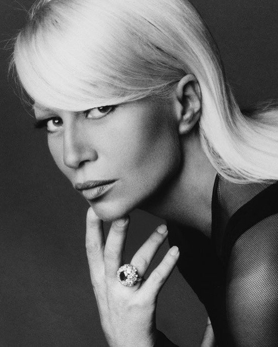 Modelo posa com expressão intensa, usando blusa de tule preta e anel Versace grande com detalhes brilhantes, em foto