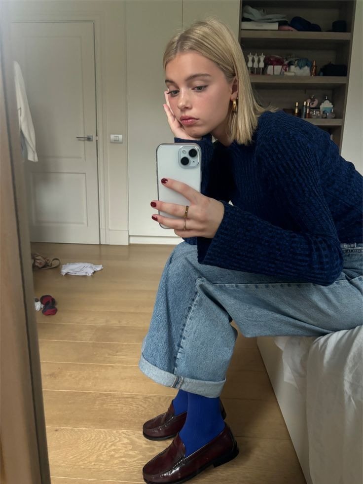 Mocassim feminino com calça jeans ampla, tricô azul e meia vibrante; modelo posa sentada no quarto com ar casual.