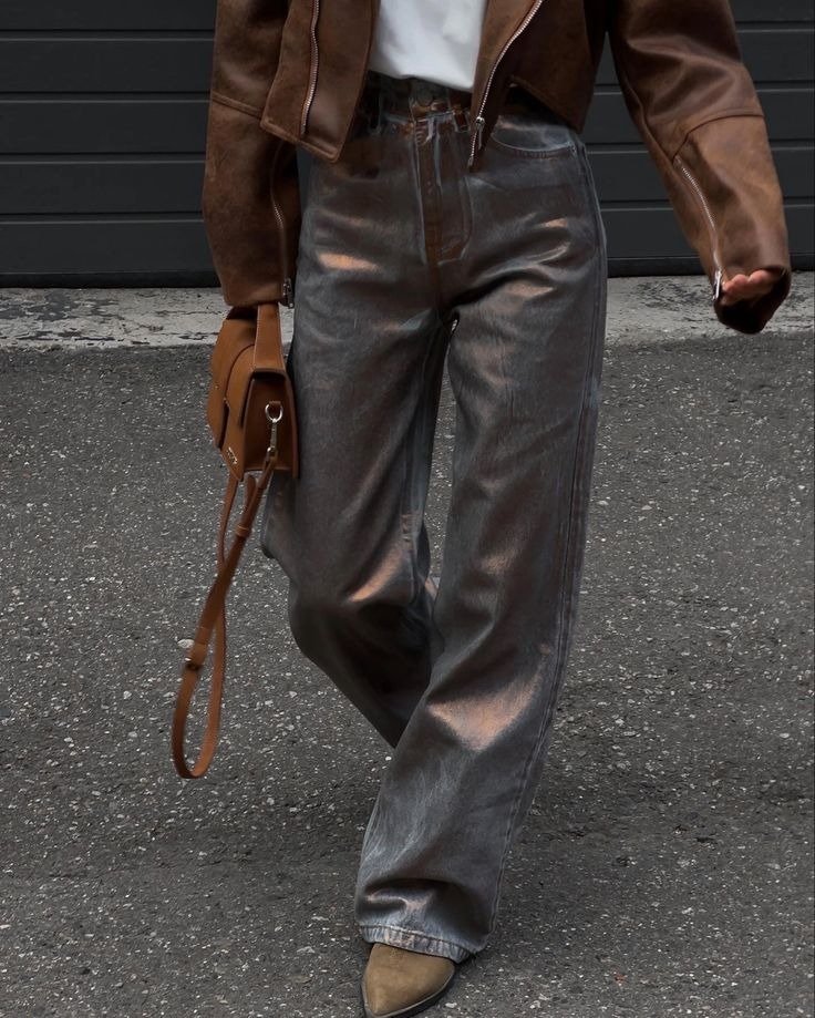 Modelo caminha com calça resinada cinza, jaqueta marrom e bolsa caramelo em cena urbana de atitude sofisticada