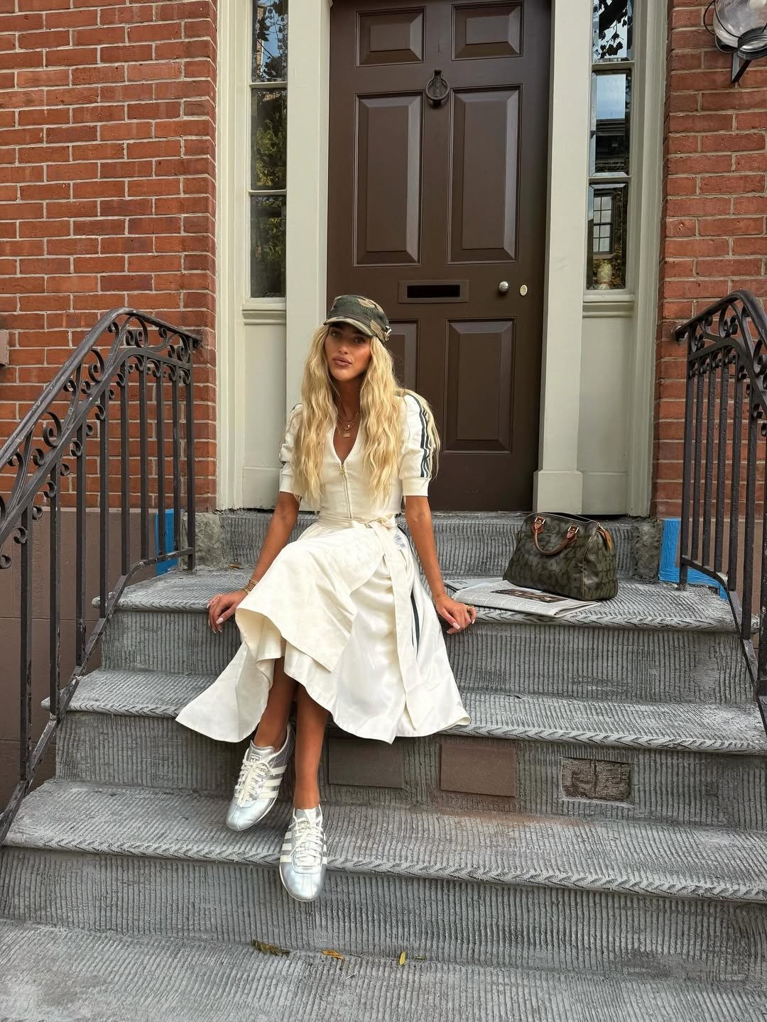 Modelo posa na escada com vestido branco fluido, boné camuflado e Tênis da Adidas baratos e estilosos prateados.