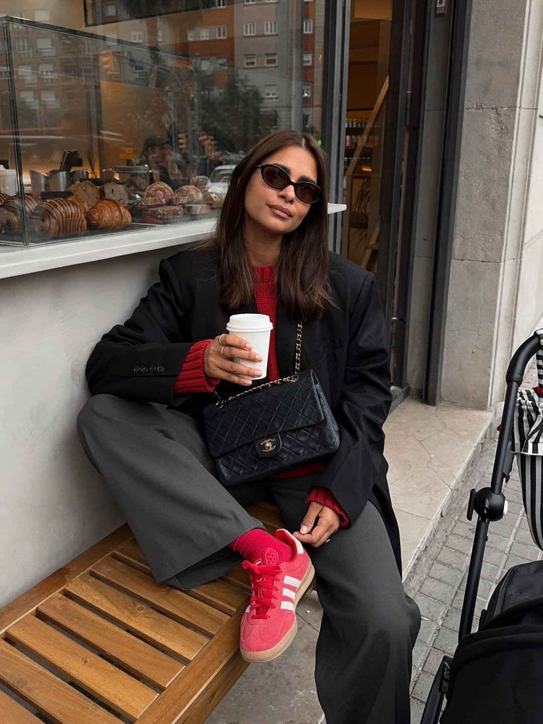 Modelo posa em café com blazer preto, tricô vermelho e Tênis da Adidas baratos e estilosos rosa, em clima urbano casual.