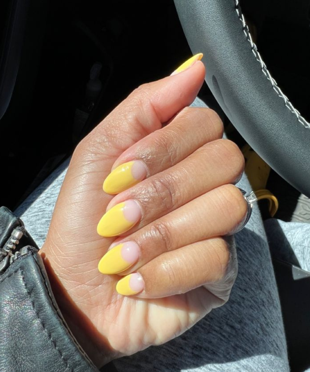 Esmaltação polêmica com unhas amareladas em formato oval, destacando a meia-lua natural, sob luz solar dentro de carro.