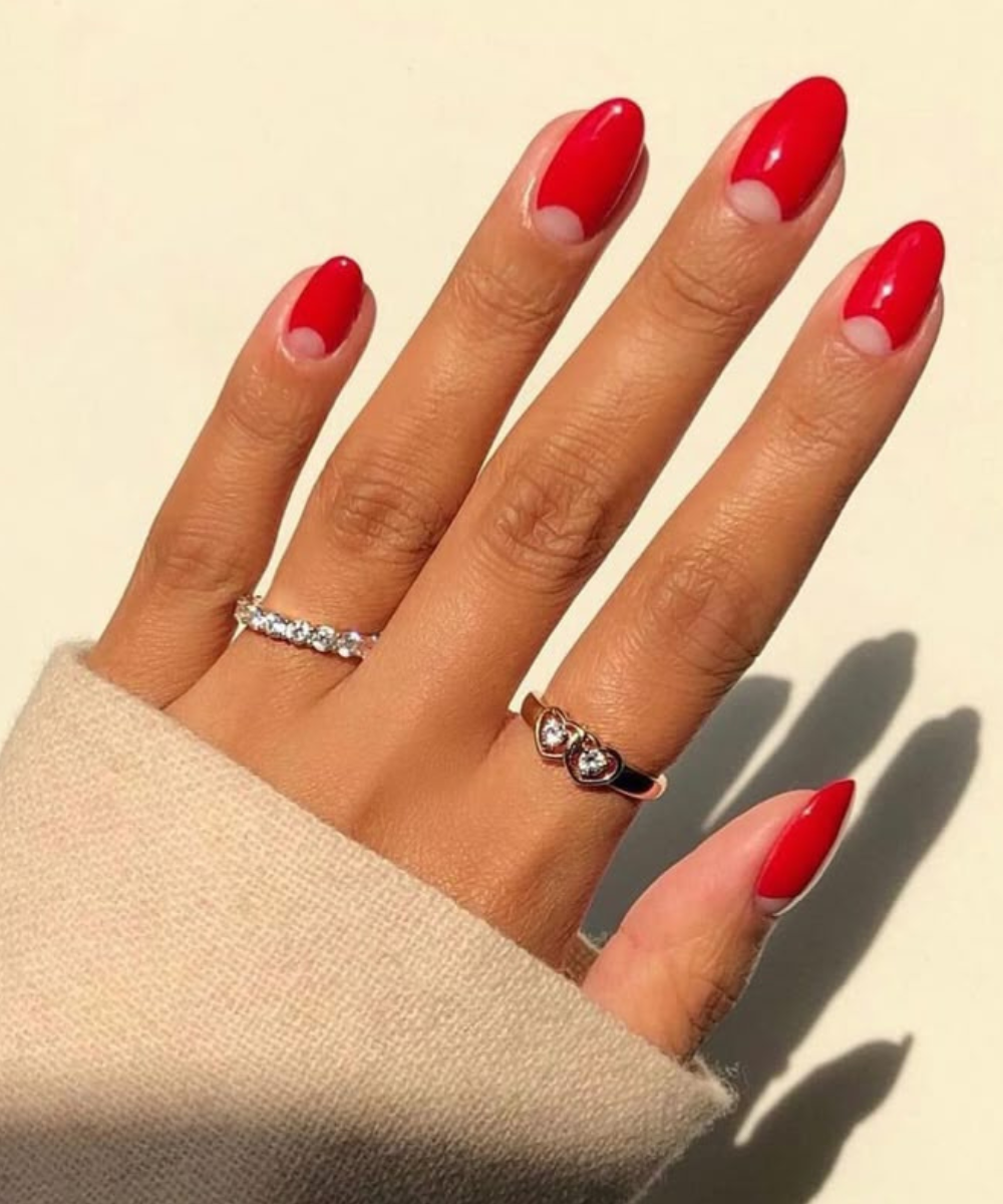 Esmaltação polêmica em unhas ovais com base natural visível e ponta vermelha vibrante, combinando anéis delicados e suéter