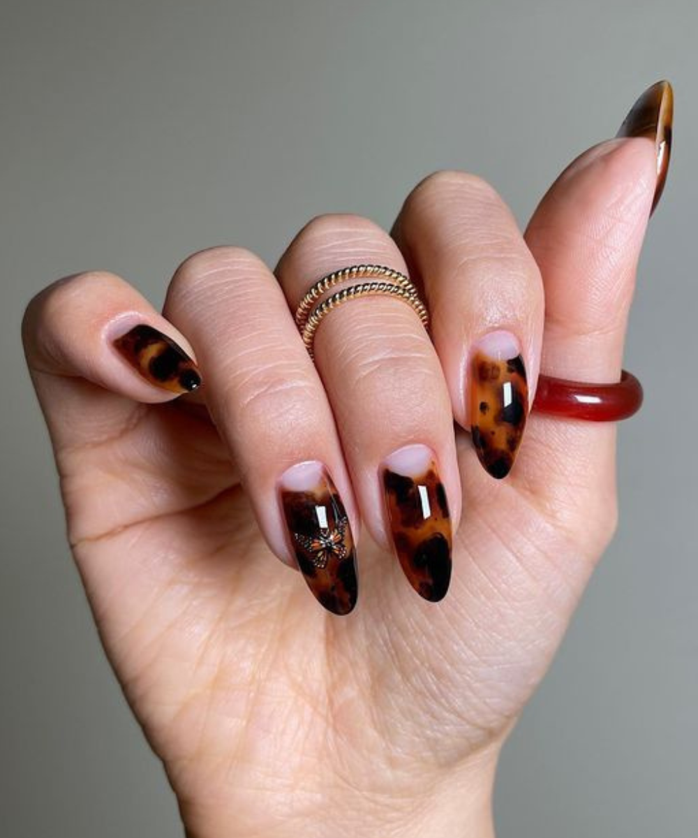 Unhas longas com esmaltação polêmica em padrão tartaruga marrom e preto brilhante, destacando anéis dourados e vermelho