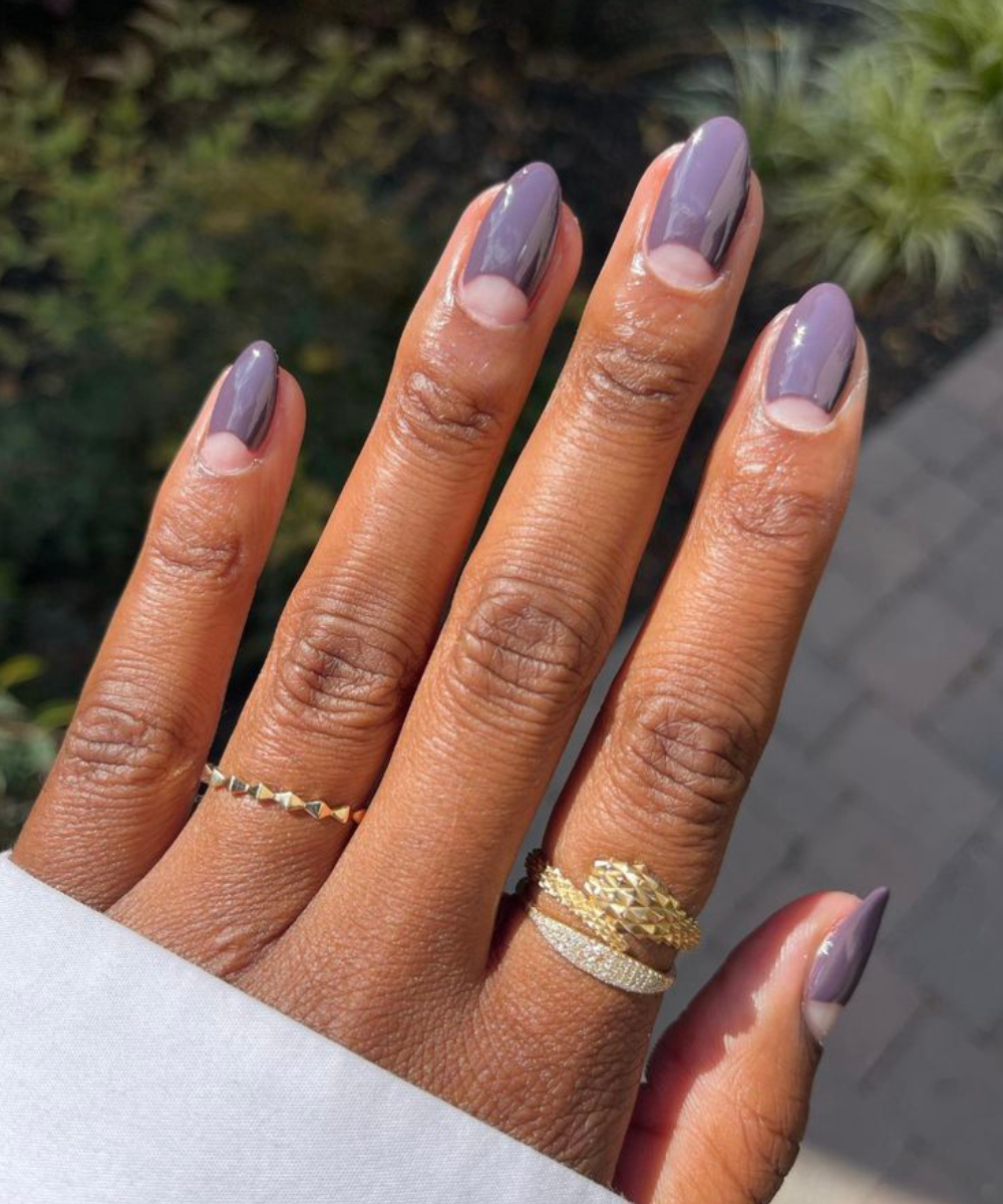Esmaltação polêmica com unhas ovais em tom lilás fosco e meia-lua natural, realçada por anéis dourados texturizados em pele