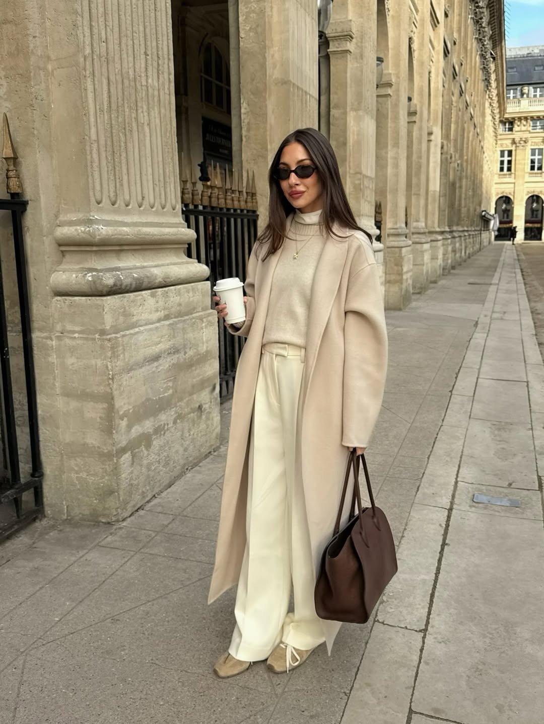 Looks com calça wide leg: modelo posa com tricô e casaco bege, tons neutros e elegância urbana entre colunas.