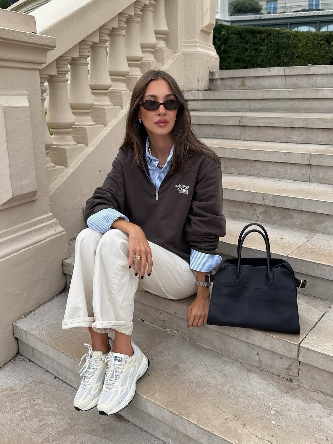 Looks com tênis esportivos: modelo posa sentada em escada de pedra com moletom marrom, camisa listrada e calça off-white.