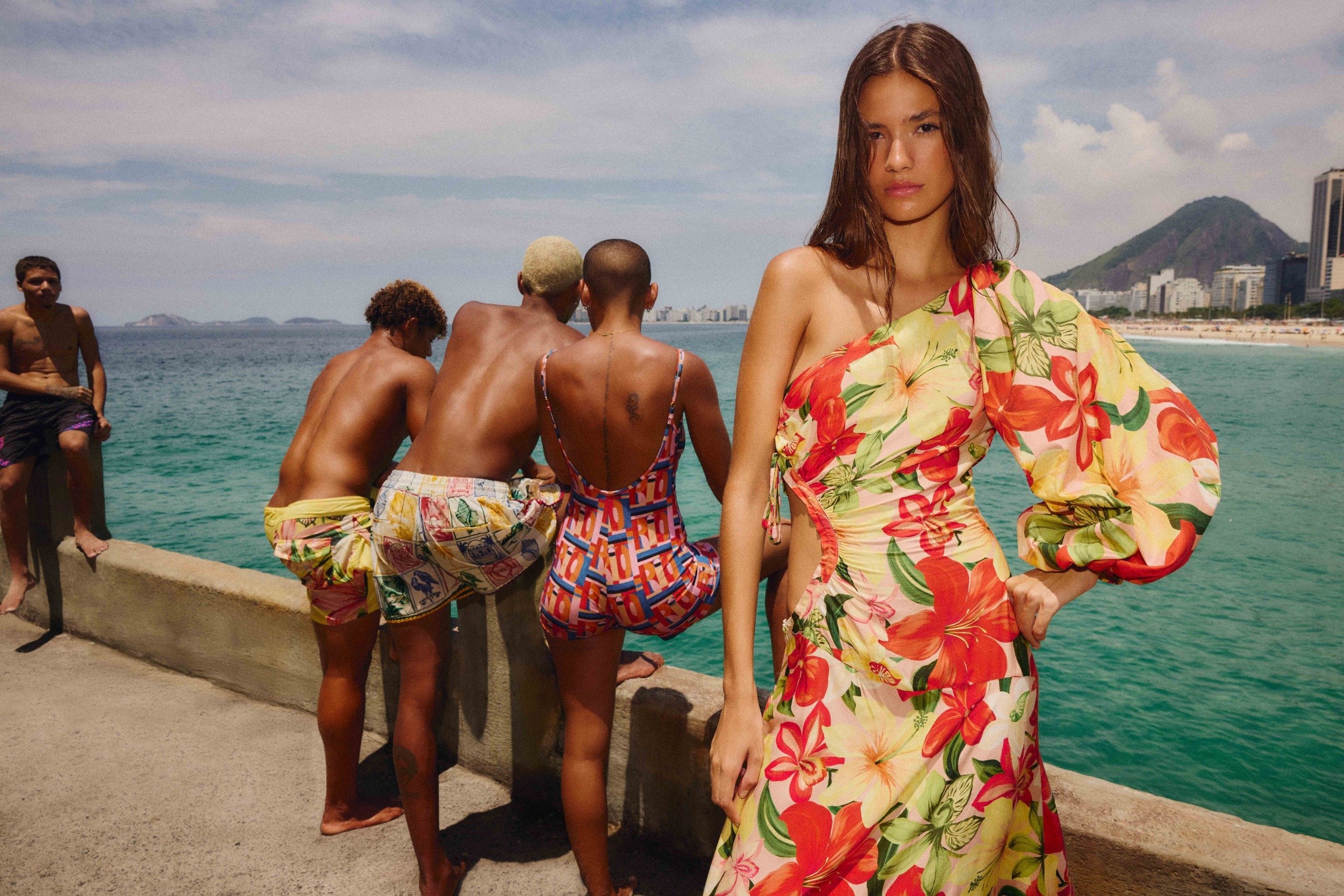 Modelo posa confiante com vestido assimétrico floral em amarelo e vermelho, enquanto grupo ao fundo veste estampas vibrantes