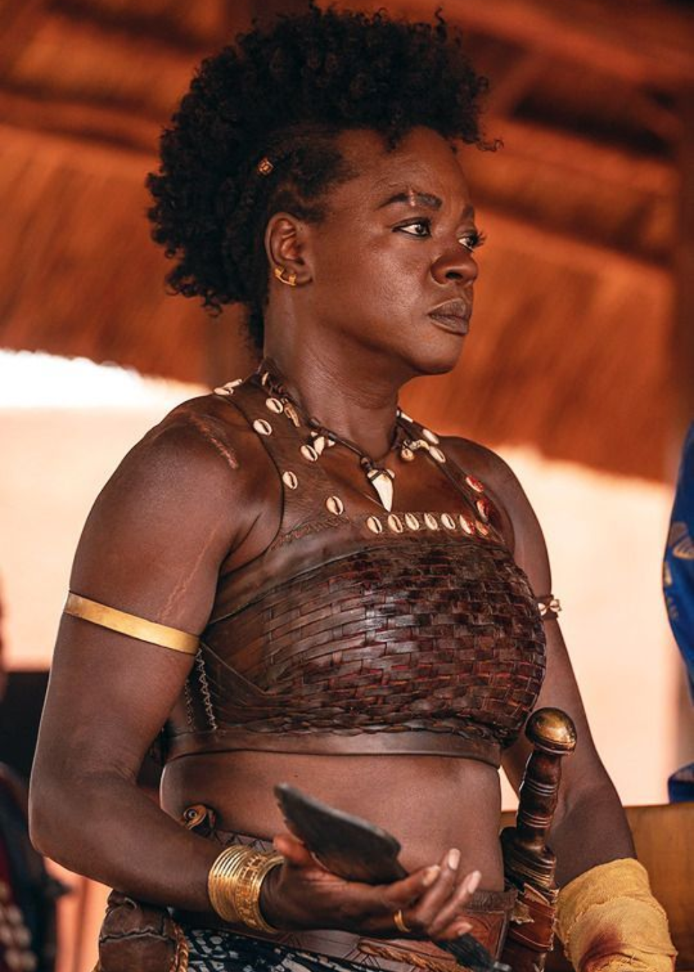 Mulher em postura firme veste top de couro trançado com búzios, braceletes dourados e segura faca.