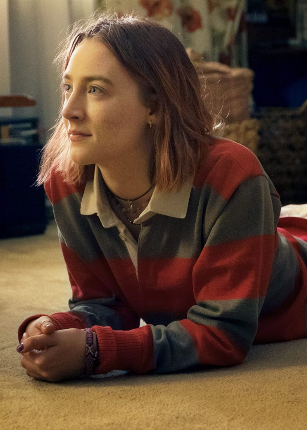 Jovem deitada no chão com suéter listrado vermelho e cinza em malha, expressão serena e olhar esperançoso.