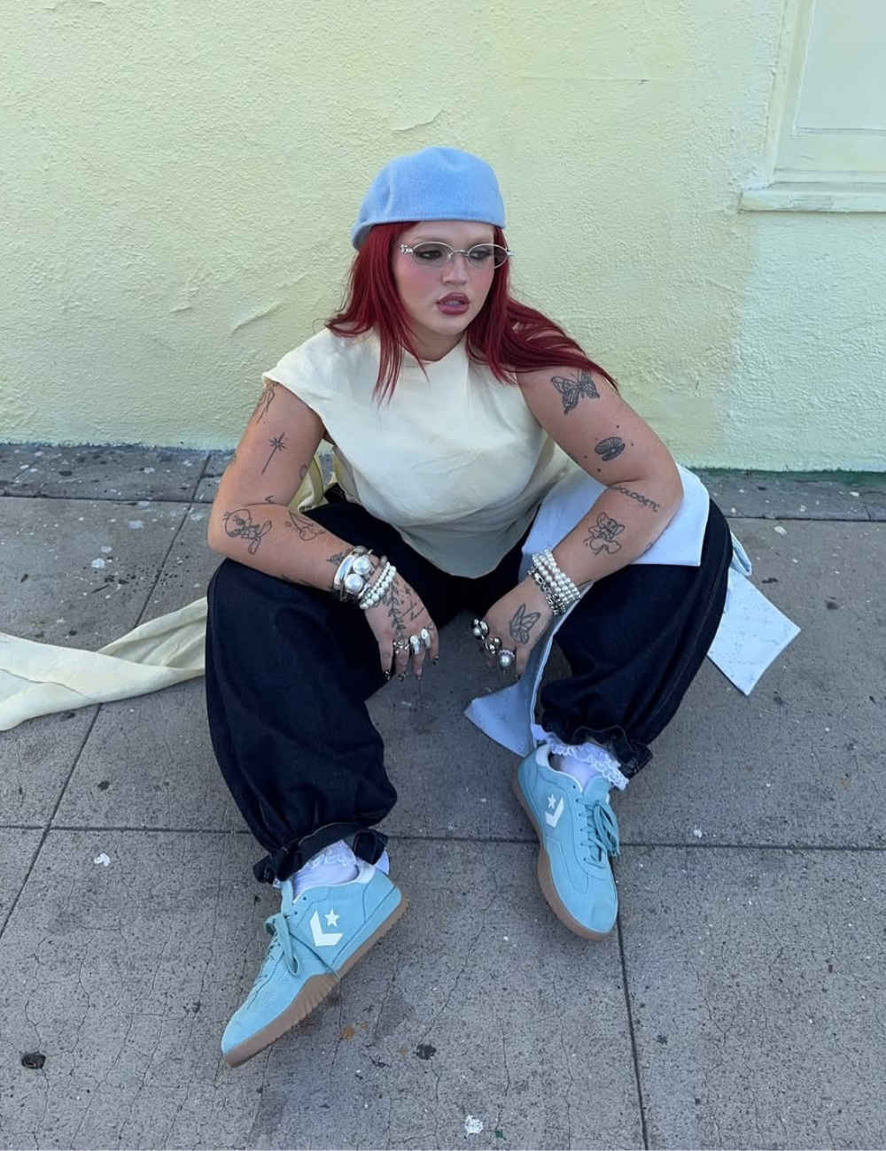 Looks de última hora para festivais: modelo agacha com boina azul, regata off-white, calça ampla escura e tênis pastel.