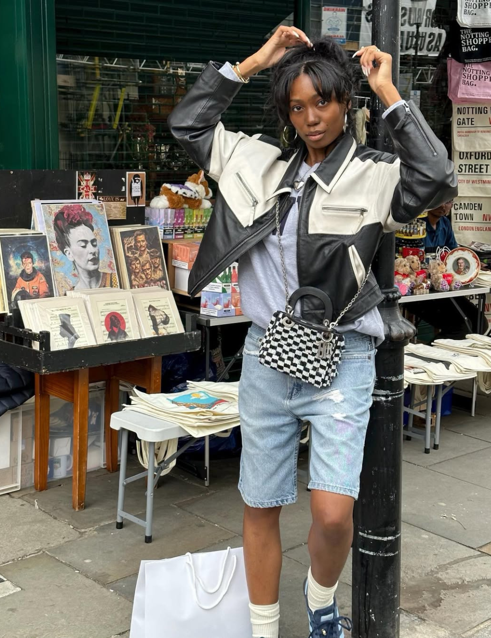 Looks de última hora para festivais: modelo posa de jaqueta bicolor, bermuda jeans e bolsa xadrez em feira urbana descolada.