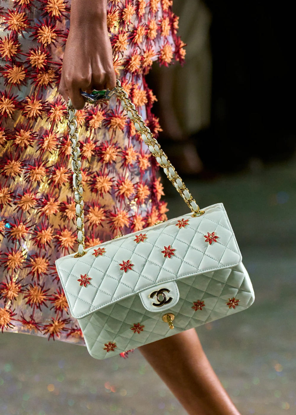 Modelo desfila com bolsa Chanel matelassê branca de corrente dourada e vestido floral texturizado em tons quentes.