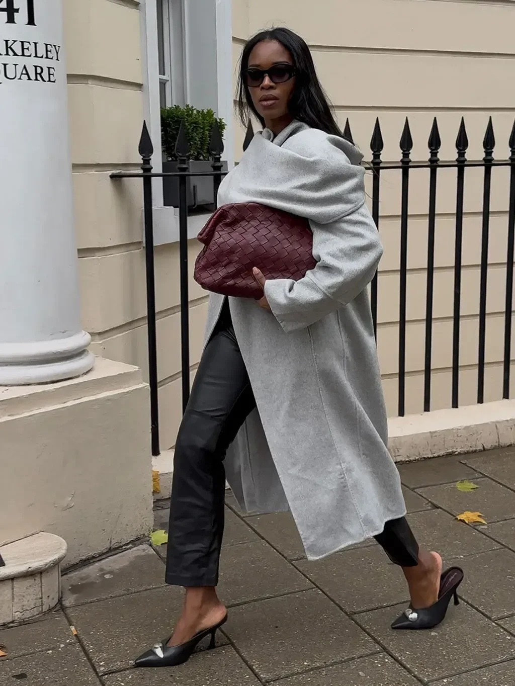 Modelo posa com calça resinada preta, casaco cinza longo e bolsa vinho em rua elegante.