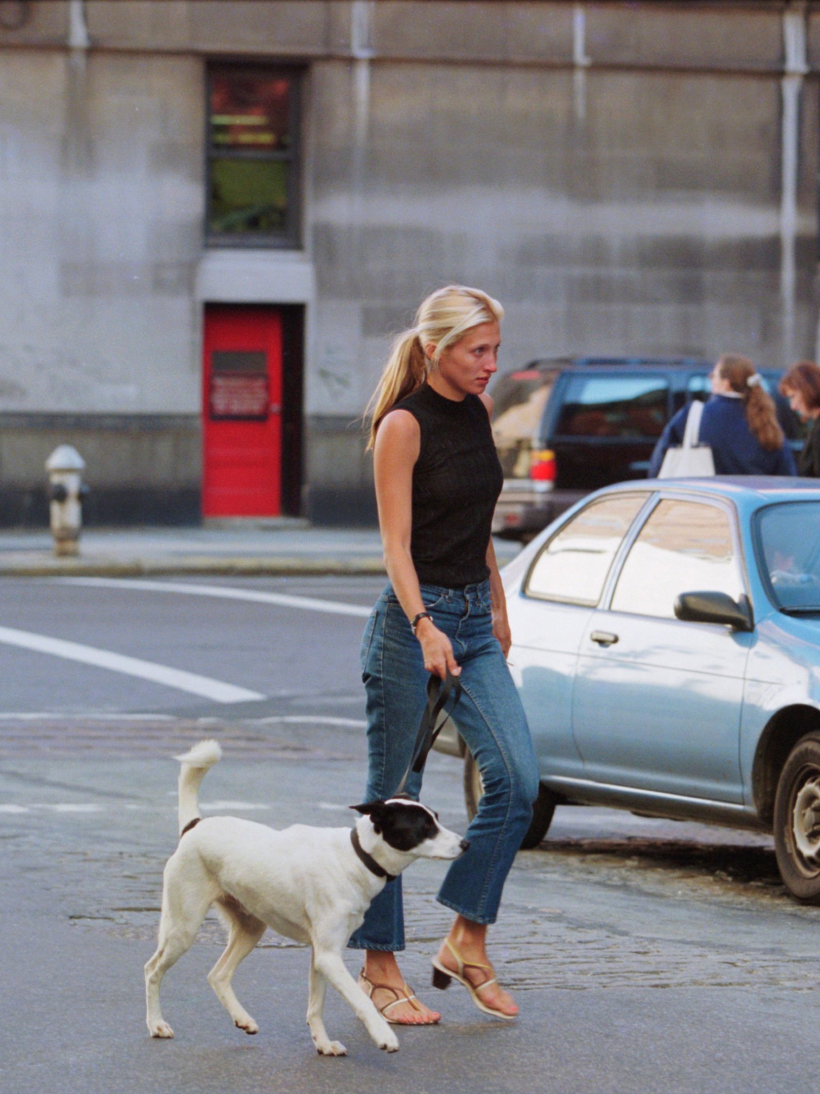Carolyn Bessette-Kennedy cruza a rua com regata preta, jeans reto e sandálias, em clima urbano minimalista.