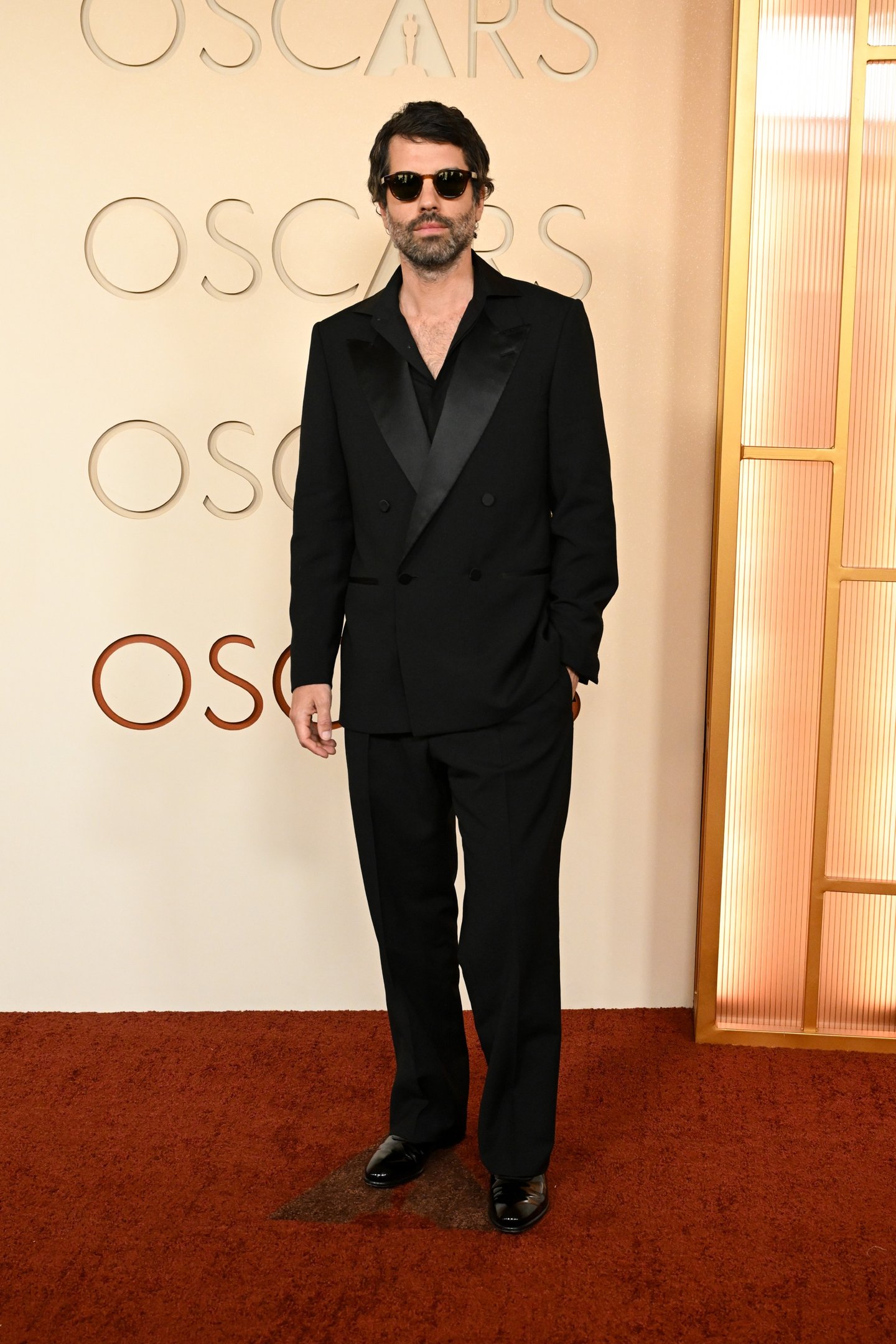 Brasileiros no Oscar: homem posa de terno preto amplo com lapela acetinada e óculos escuros no tapete vermelho.