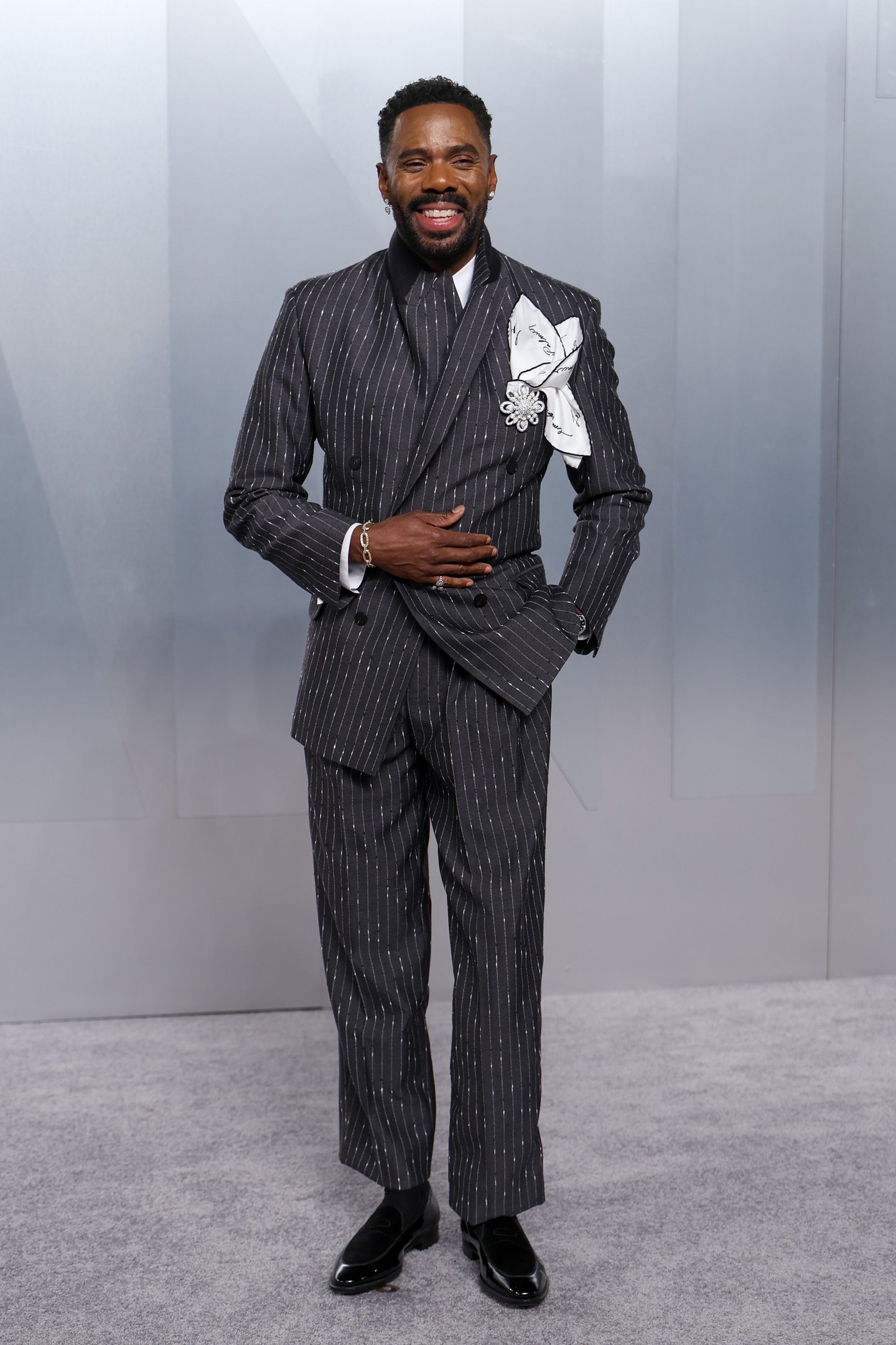 After party do Oscar 2026: modelo posa sorrindo em terno risca de giz cinza com flor branca, elegância festiva.