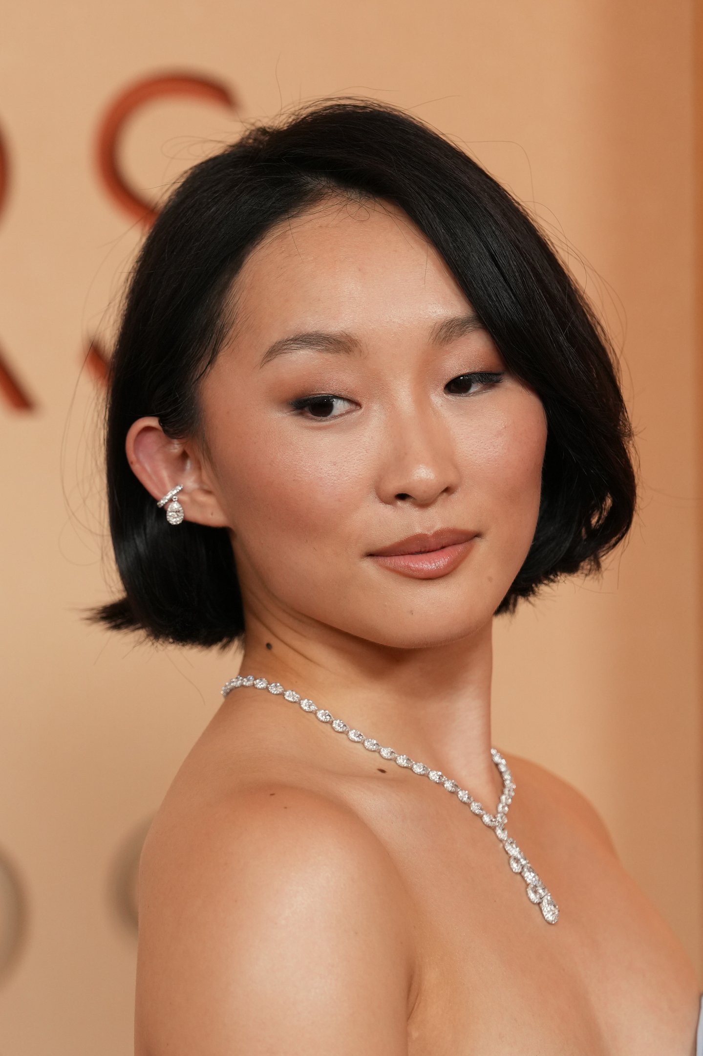 Modelo posa com batom nude rosado, bob preto liso e joias brilhantes sobre pele iluminada em fundo bege elegante.