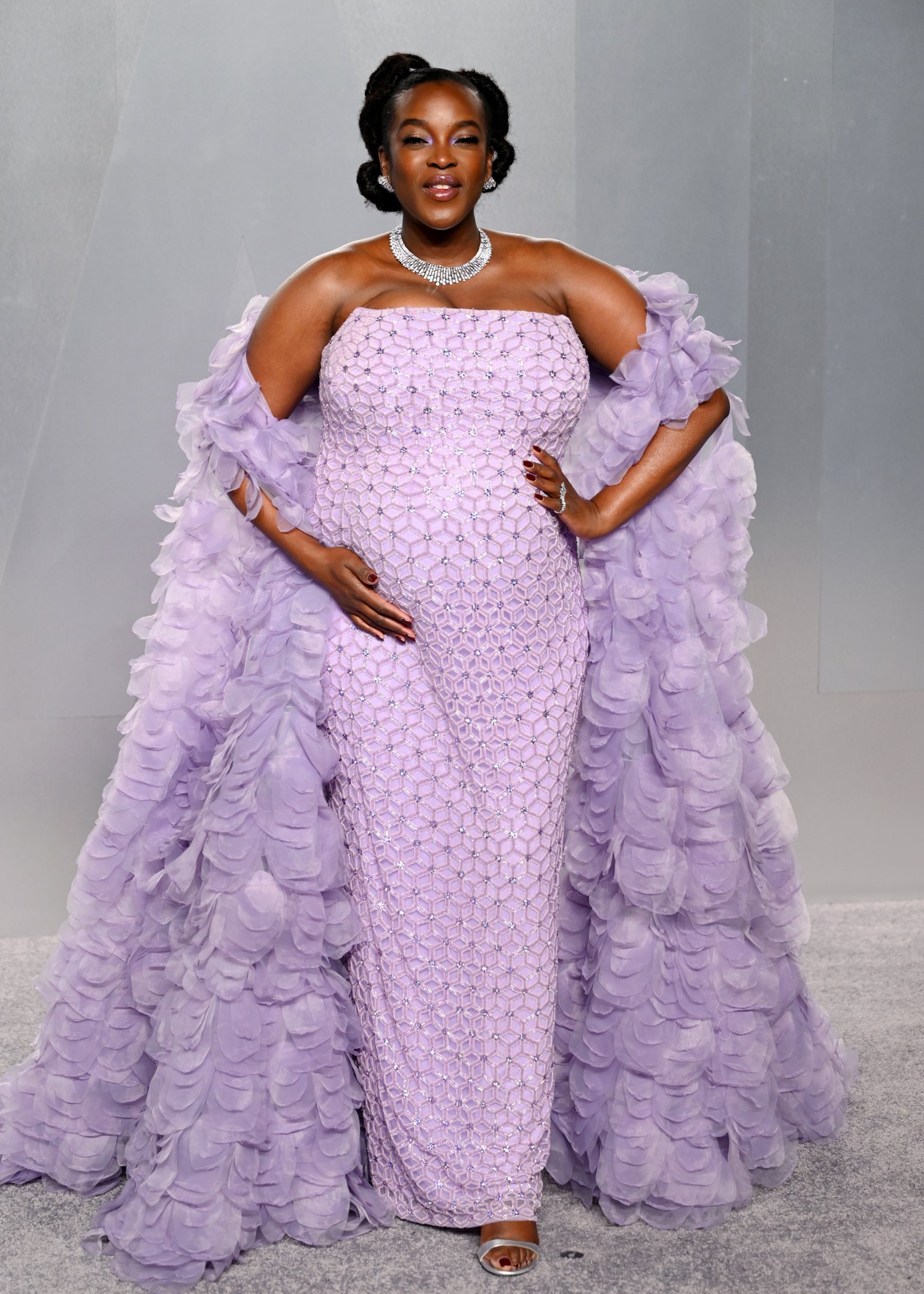 After party do Oscar 2026: modelo posa com vestido lilás tomara que caia bordado e capa de babados, em clima glamouroso.