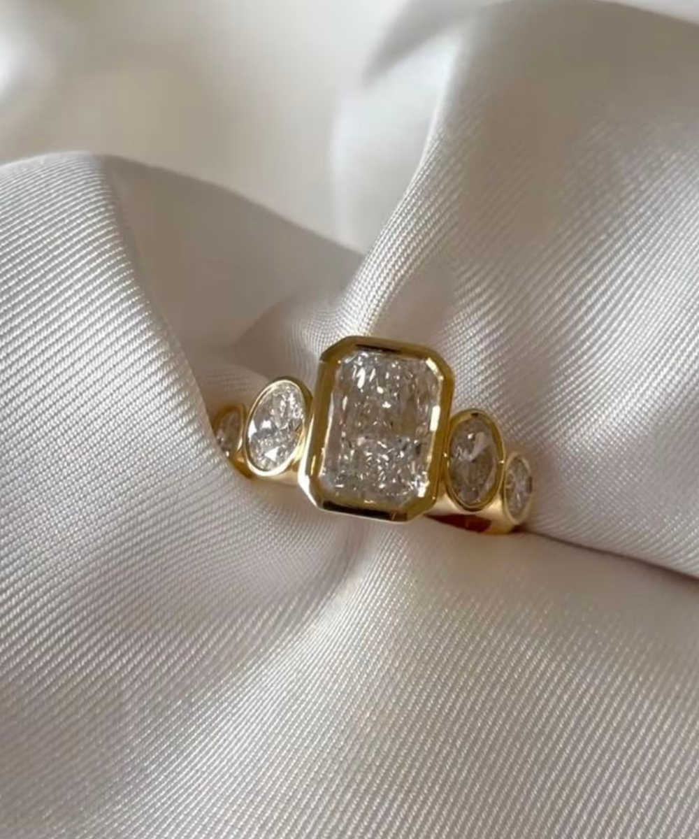 Anel de noivado em ouro amarelo com diamante central quadrado e pedras laterais ovais, sobre tecido branco acetinado,