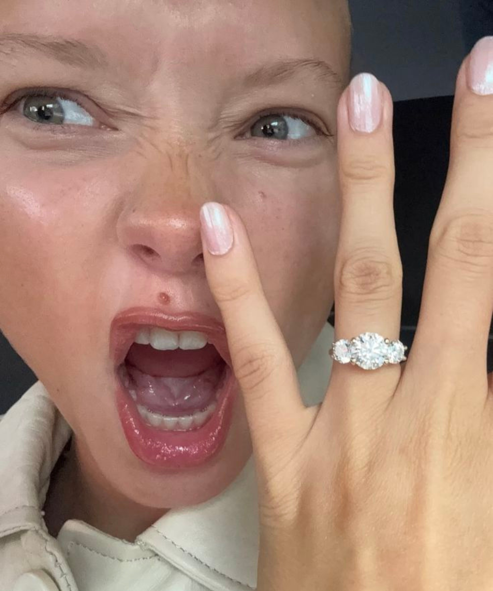 Mulher exibe anéis de noivado com pedras brilhantes em prata, expressando surpresa e alegria com esmalte rosa claro.