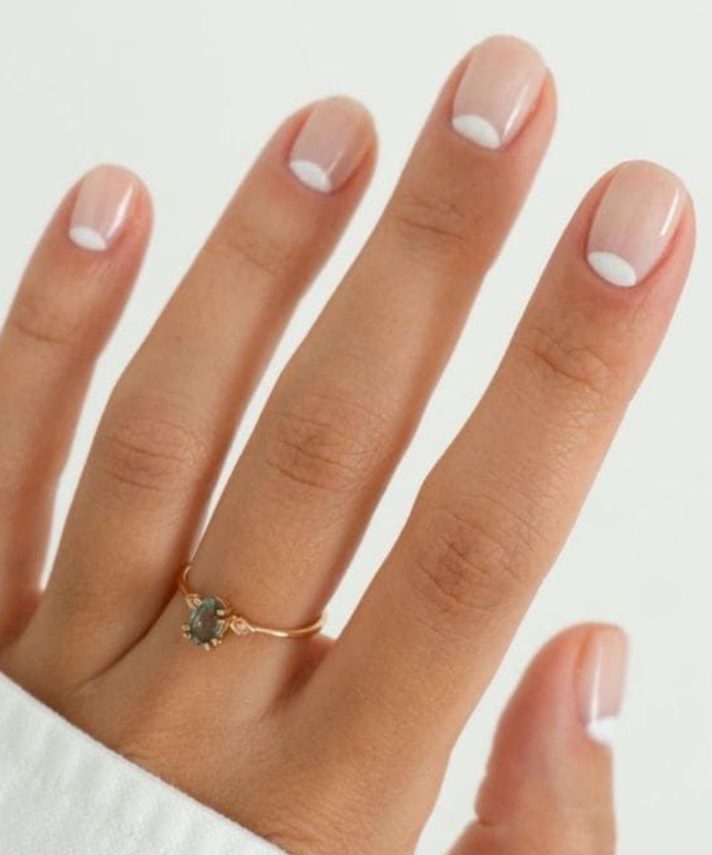 Unhas com esmaltação polêmica em tom nude com meia-lua branca na base, destacando anel delicado dourado com pedra cinza.