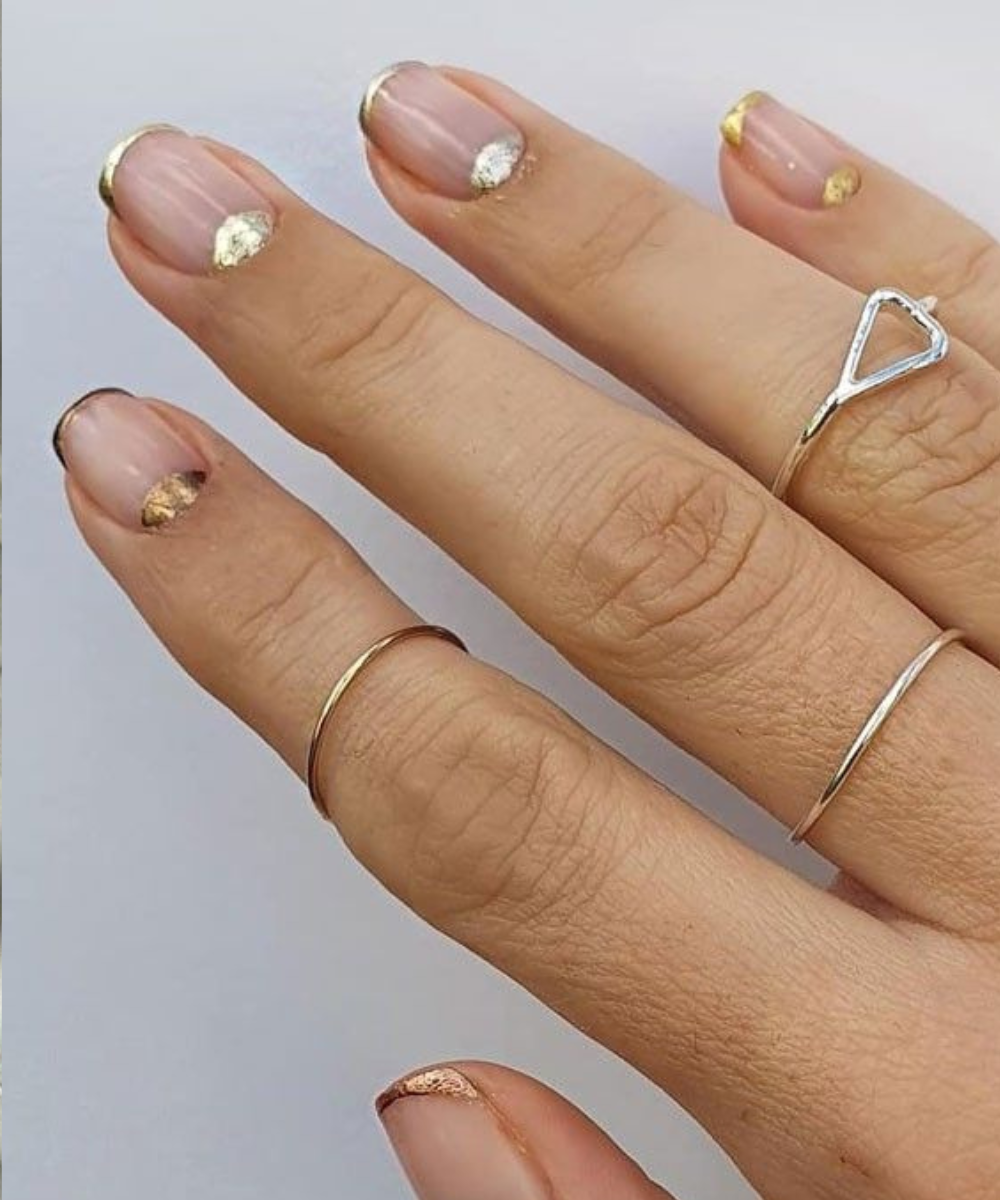 Unhas curtas com esmaltação polêmica em base nude e detalhes assimétricos em dourado metálico, combinando com anéis finos em