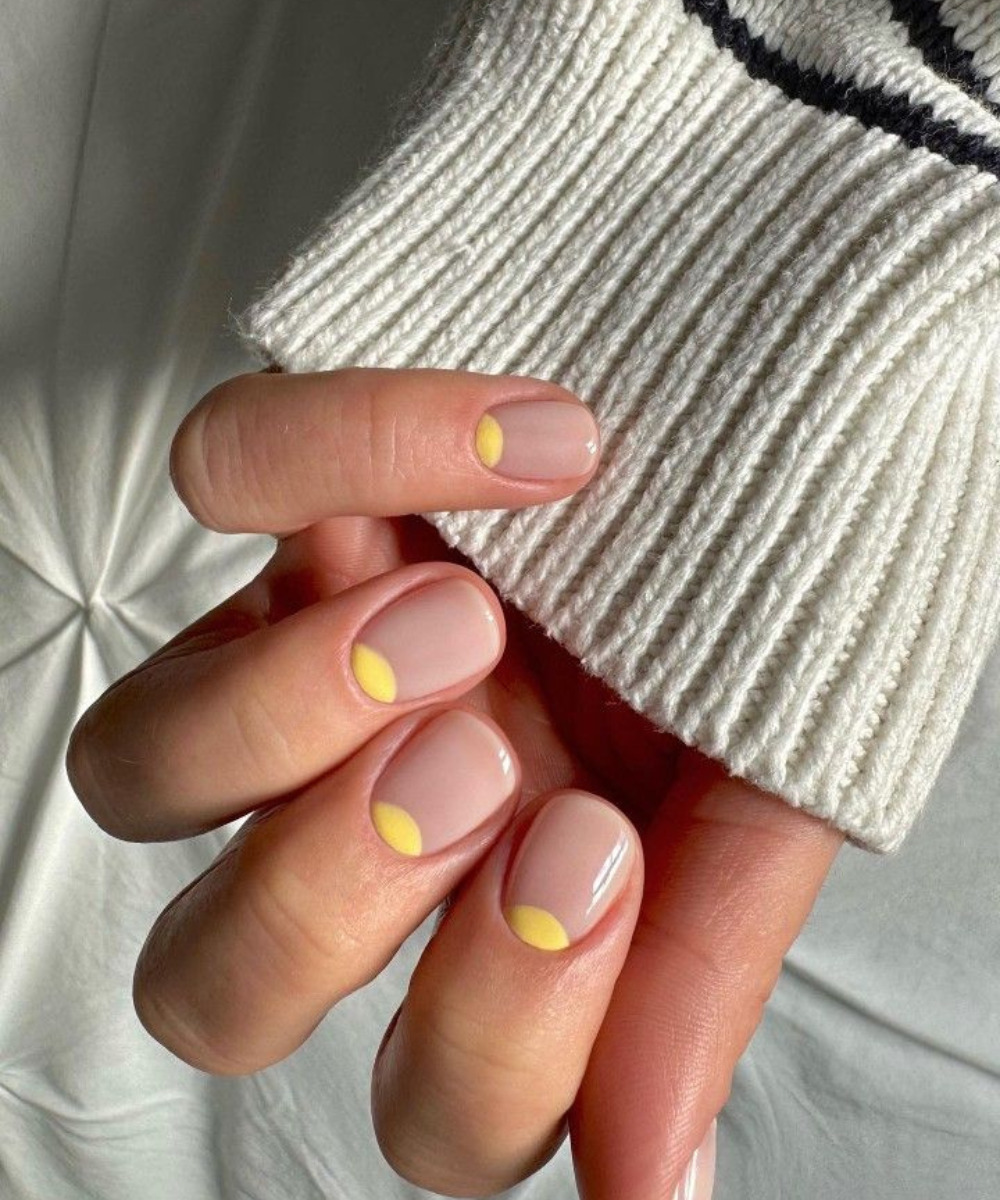 Esmaltação polêmica com base nude acetinada e meia-lua amarela vibrante destaca unhas curtas e arredondadas em clima